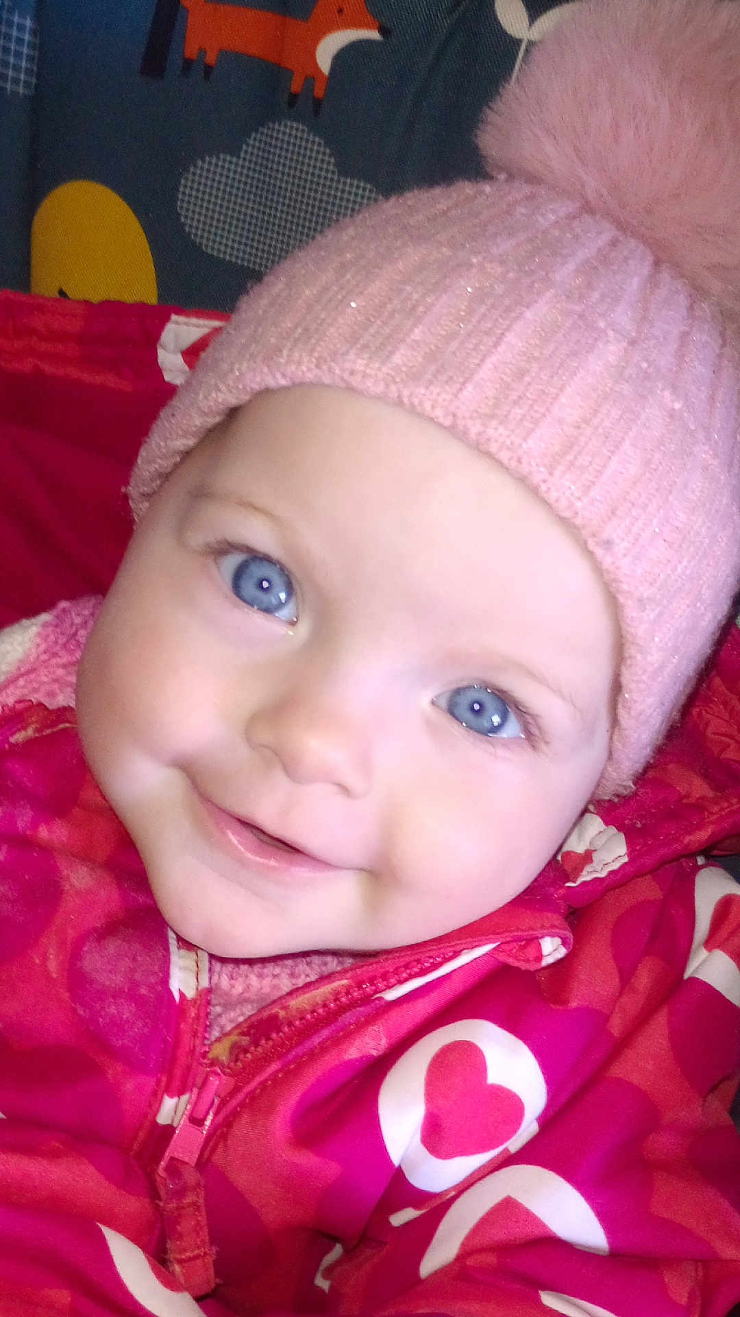 Chelsea is registered to the contest to win money with this photo: baby, child, blue_eyes, pink_hat, pom_pom, knit_hat, jacket, heart_pattern, smile, close_up, face, cute, warm_clothing, infant, portrait, cozy, colorful, indoor, happy, young
