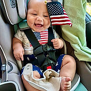 Toby joined the competition — help win amazing prizes! baby, car_seat, american_flag, smiling, child, infant, safety_harness, clothing, blue_shorts, white_shirt, stars, stripes, cute, happy, indoors, car_interior, seatbelt, toddler, flag, hand