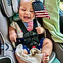 baby, car_seat, american_flag, smiling, child, infant, safety_harness, clothing, blue_shorts, white_shirt, stars, stripes, cute, happy, indoors, car_interior, seatbelt, toddler, flag, hand
