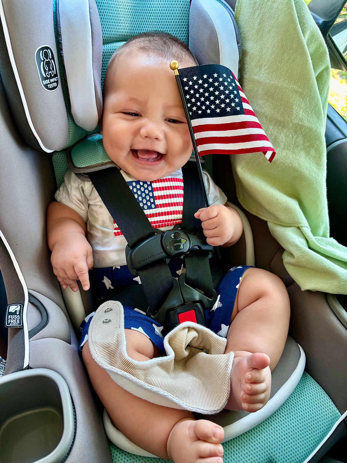 Toby joined the competition — help win amazing prizes! baby, car_seat, american_flag, smiling, child, infant, safety_harness, clothing, blue_shorts, white_shirt, stars, stripes, cute, happy, indoors, car_interior, seatbelt, toddler, flag, hand