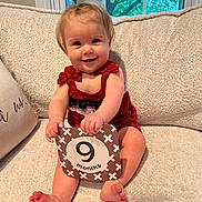 Magnolia is registered to the contest to win money with this photo: baby, child, infant, red_clothing, smiling, sitting, milestone_card, 9_months, indoors, couch, window, greenery, hair_tie, skin, feet, hands, happy, portrait, cute, home