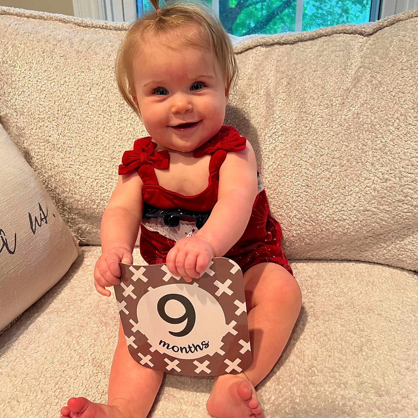 Magnolia is registered to the contest to win money with this photo: baby, child, infant, red_clothing, smiling, sitting, milestone_card, 9_months, indoors, couch, window, greenery, hair_tie, skin, feet, hands, happy, portrait, cute, home