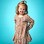 child, girl, smile, posing, dress, checkered_pattern, curly_hair, topknot, studio, blue_background, casual_clothing, happy, young, portrait, fashion, standing, playful, confident, long_sleeves, buttoned_dress