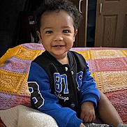 Aziah joined the competition — help win amazing prizes! toddler, child, smiling, curly_hair, jacket, blue, black, blanket, crocheted_blanket, indoor, bedroom, socks, happy, person, portrait, seated, furniture, door, home, cute