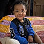 toddler, child, smiling, curly_hair, jacket, blue, black, blanket, crocheted_blanket, indoor, bedroom, socks, happy, person, portrait, seated, furniture, door, home, cute