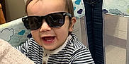 Alexander is registered to the contest to win money with this photo: child, baby, toddler, sunglasses, striped_shirt, playpen, hand, ring, smile, teeth, indoor, filing_cabinet, cabinet, mesh, blanket, person, portrait, happy, clothing, nursery