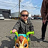 adult, asphalt, car, child, cute, daylight, fun, jacket, outdoor, parking_lot, person, play, shoes, smile, sunglasses, toddler, tongue_out, toy, toy_horse, vehicle