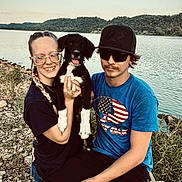 Lanna is registered to the contest to win money with this photo: lake, water, dog, puppy, black_and_white, person, glasses, hat, blue_shirt, outdoor, nature, rocks, grass, smiling, casual_clothing, young_adult, sitting, sunlight, landscape, friendship
