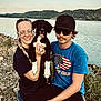 lake, water, dog, puppy, black_and_white, person, glasses, hat, blue_shirt, outdoor, nature, rocks, grass, smiling, casual_clothing, young_adult, sitting, sunlight, landscape, friendship