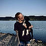 Lanna is registered to the contest to win money with this photo: woman, dog, puppy, lake, water, rocks, outdoor, nature, sky, casual_clothing, glasses, braids, affection, pet, holding, jeans, tattoo, sunlight, portrait, smiling