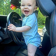 Julien is registered to the contest to win money with this photo: baby, child, car, steering_wheel, smile, happy, blue_clothing, seat, interior, window, greenery, sunlight, hands, head, face, eyes, ears, toy, colorful, vehicle