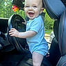 baby, blue_clothing, car, child, colorful, ears, eyes, face, greenery, hands, happy, head, interior, seat, smile, steering_wheel, sunlight, toy, vehicle, window