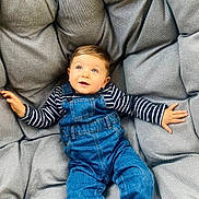 Jaïro participe au concours pour gagner de l'argent avec cette photo : toddler, child, denim_overalls, striped_shirt, cushion, gray_cushion, plush, comfortable, indoor, casual_clothing, cute, portrait, baby, smiling, happy, relaxed, young_child, person, seated, blue_eyes