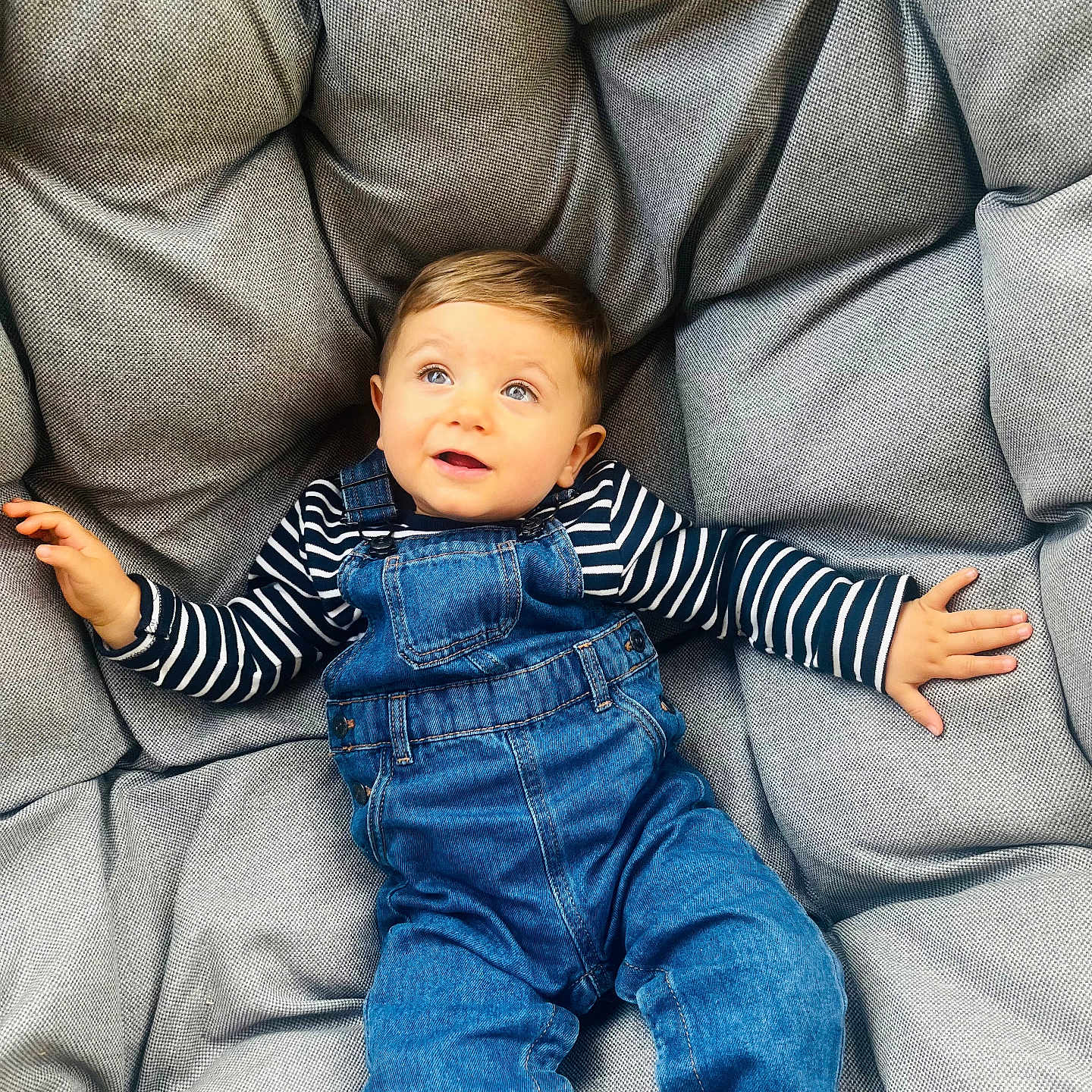 Jaïro participe au concours pour gagner de l'argent avec cette photo : baby, blue_eyes, casual_clothing, child, comfortable, cushion, cute, denim_overalls, gray_cushion, happy, indoor, person, plush, portrait, relaxed, seated, smiling, striped_shirt, toddler, young_child