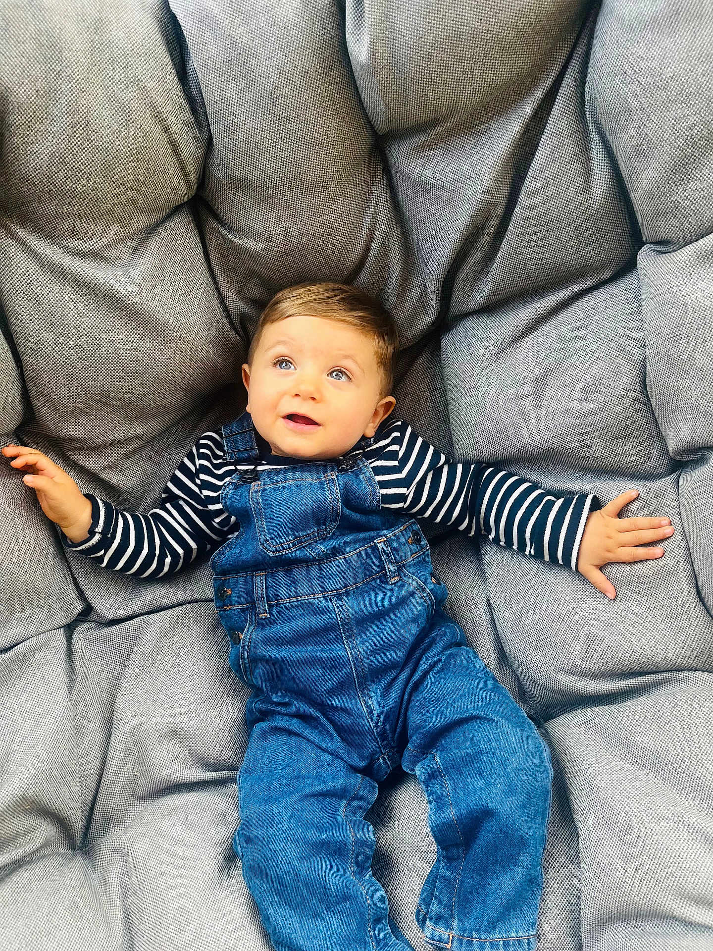 Jaïro participe au concours pour gagner de l'argent avec cette photo : toddler, child, denim_overalls, striped_shirt, cushion, gray_cushion, plush, comfortable, indoor, casual_clothing, cute, portrait, baby, smiling, happy, relaxed, young_child, person, seated, blue_eyes