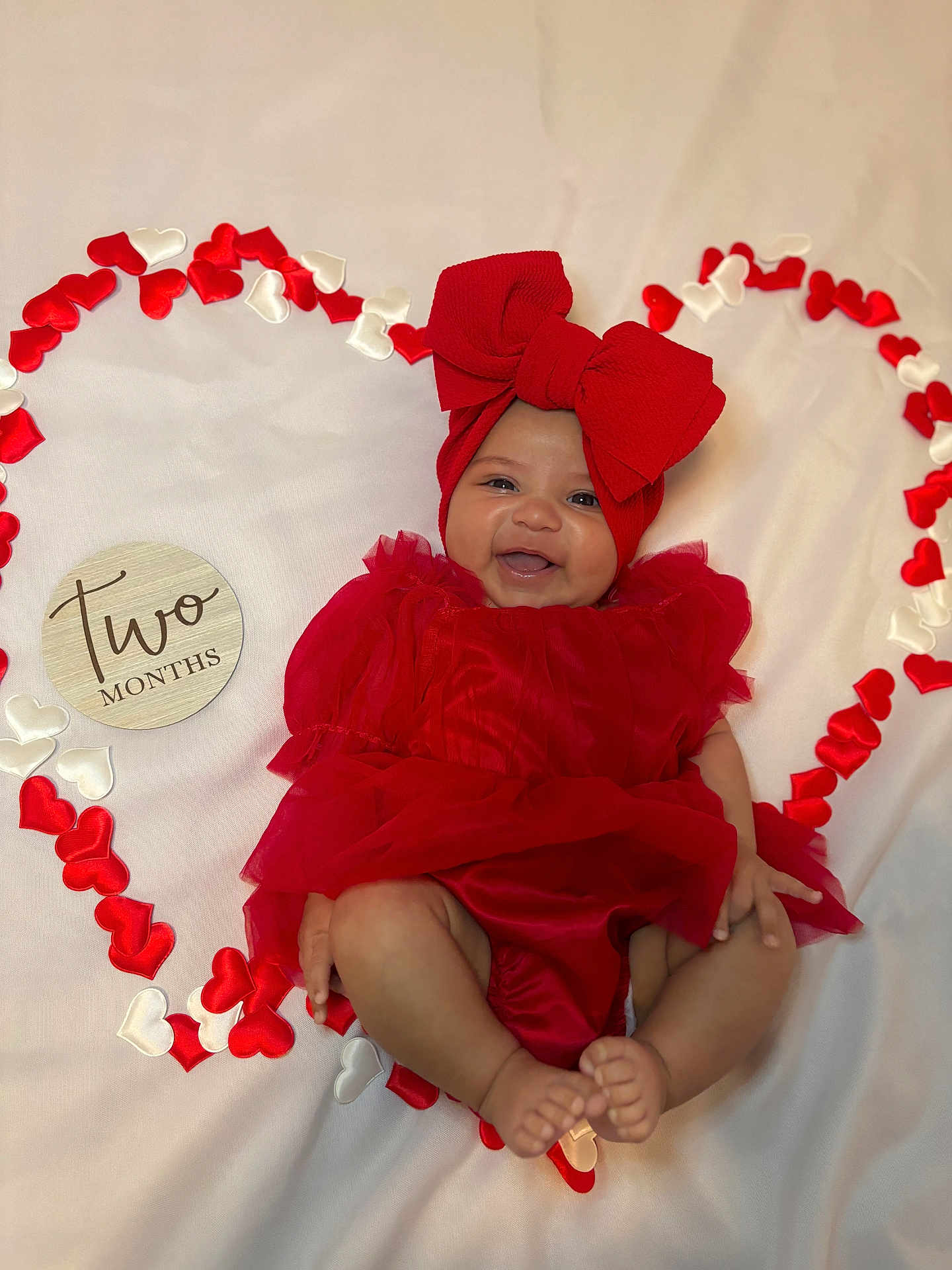 Araceli is registered to the contest to win money with this photo: baby, infant, smiling, red_dress, headband, bow, heart_shape, fabric, white_background, celebration, cute, happy, two_months, portrait, laying_down, skin, feet, hands, closeup, decorative_hearts