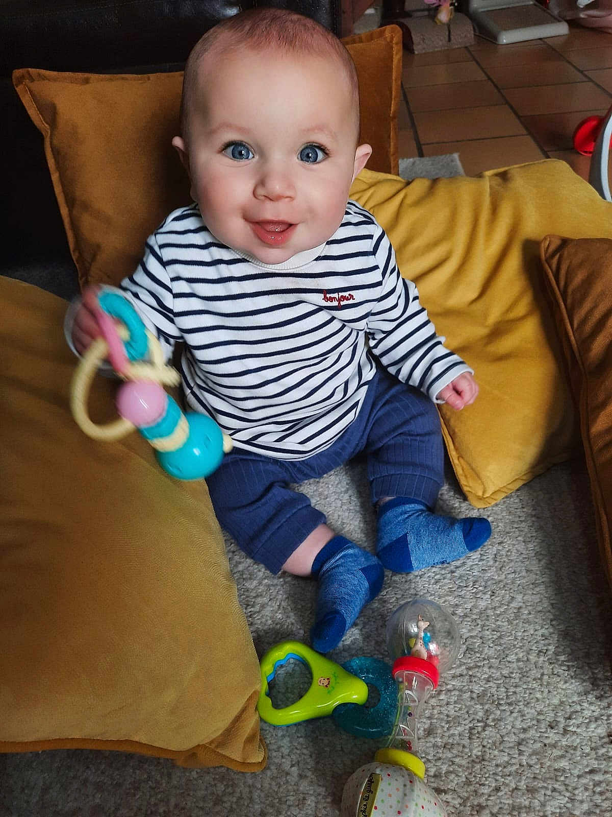 Elio participe au concours pour gagner de l'argent avec cette photo : baby, child, toy, blue_eyes, striped_shirt, socks, pillow, carpet, smiling, indoor, cute, seated, playful, colorful_toy, happy, infant, home, floor, person, portrait