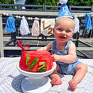 Brooks joined the competition — help win amazing prizes! baby, smiling, party_hat, watermelon, cake, outdoor, blanket, denim_overalls, celebration, half_birthday, tassel_garland, banner, sunlight, grass, foot, hands, face, happy, summer, table