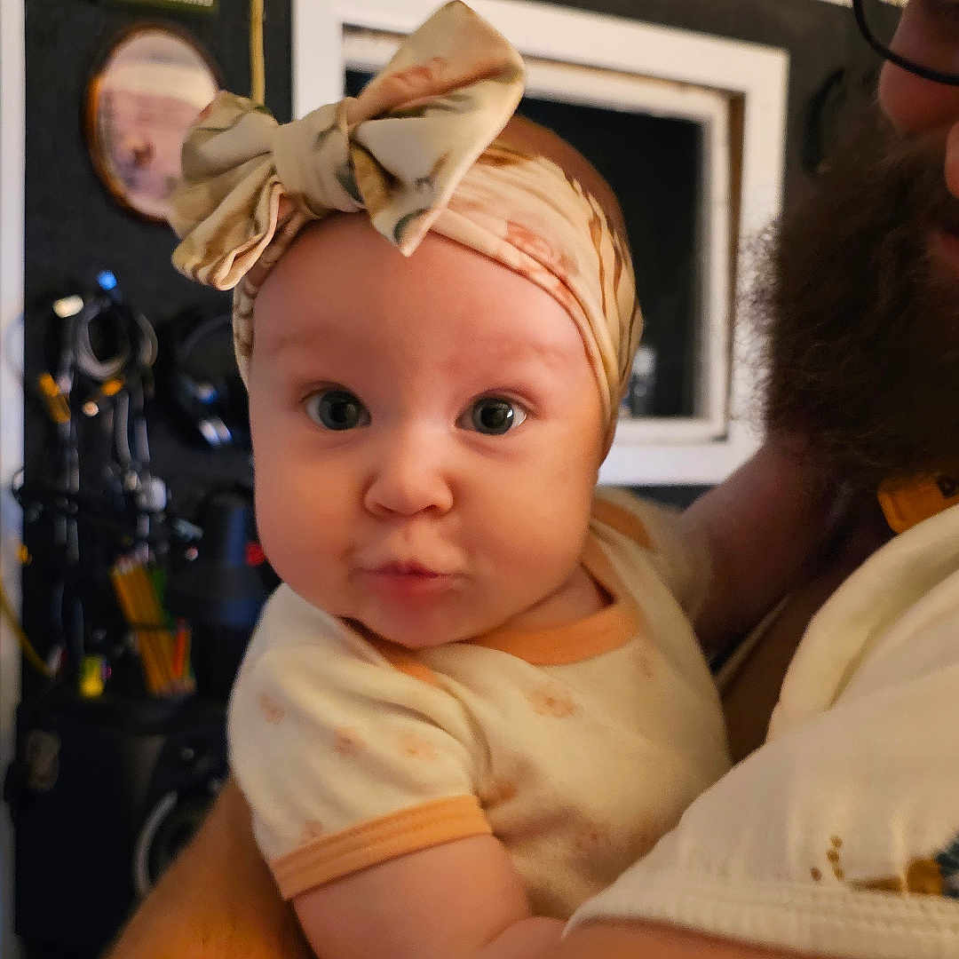 Sunni is registered to the contest to win money with this photo: accessories, baby, bonnet, cap, clothing, couch, dress, face, fireplace, furniture, glasses, hat, head, headband, indoors, person, photography, portrait, strap, wood