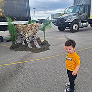 Kado is registered to the contest to win money with this photo: toddler, child, yellow_shirt, leopard, stuffed_animal, truck, parking_lot, outdoor, curious, confused, animal_graphics, plant, pavement, cloudy_sky, casual_clothing, sneakers, toy, exhibit, realistic, event