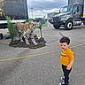 toddler, child, yellow_shirt, leopard, stuffed_animal, truck, parking_lot, outdoor, curious, confused, animal_graphics, plant, pavement, cloudy_sky, casual_clothing, sneakers, toy, exhibit, realistic, event