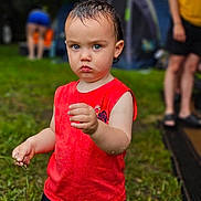 Kado is registered to the contest to win money with this photo: child, toddler, red_shirt, grass, outdoor, camping, tent, person, summer, nature, serious_expression, holding_object, wet_hair, casual_clothing, blurred_background, adult, footwear, greenery, daytime, recreation