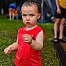 child, toddler, red_shirt, grass, outdoor, camping, tent, person, summer, nature, serious_expression, holding_object, wet_hair, casual_clothing, blurred_background, adult, footwear, greenery, daytime, recreation