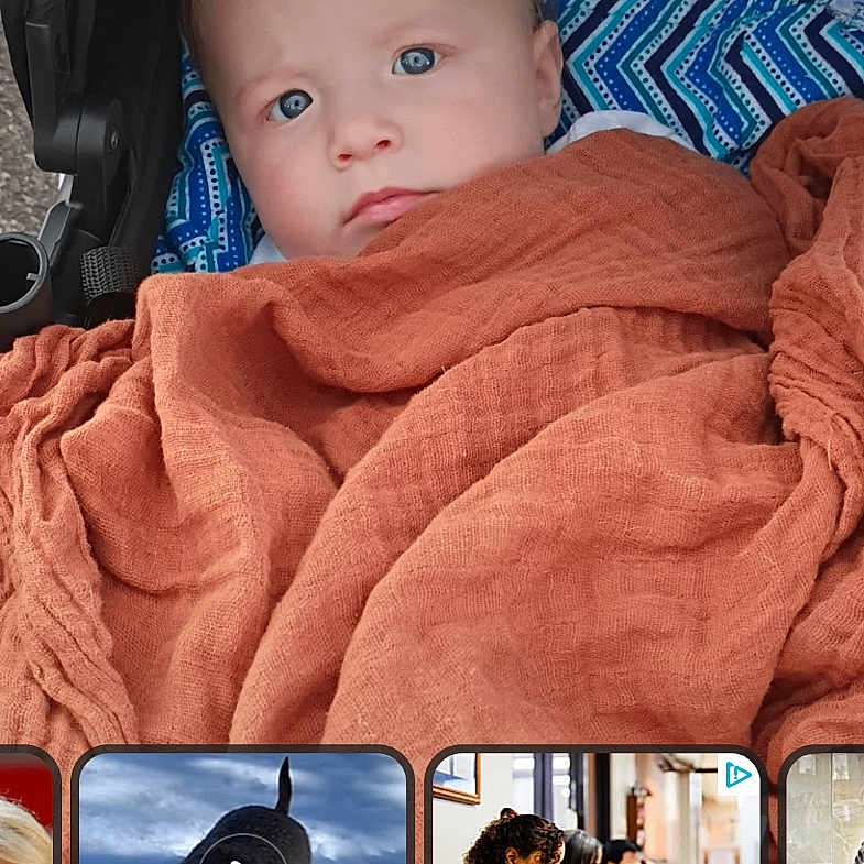 Giovanni is registered to the contest to win money with this photo: baby, infant, child, portrait, face, blue_eyes, wide_eyed, blanket, swaddled, rust_colored, stroller, seated, indoors, close_up, photograph, lock_screen, smartphone_screenshot, news_cards, cozy, adorable