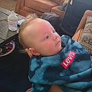 Giovanni is registered to the contest to win money with this photo: baby, child, infant, portrait, face, eyes, looking_up, blond_hair, smiling, blue_clothing, hoodie, levis, bottle, living_room, couch, coffee_table, backpack, tattoo, adult_arm, indoor