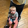 toddler, child, stroller, indoor, wooden_floor, plaid_shorts, gray_shirt, barefoot, serious_expression, blue_eyes, light_hair, baby, seatbelt, casual_clothing, small_wheels, metal_frame, floor_tiles, looking_up, hands_clenched, cute