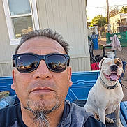 Bella Soto is registered to the contest to win money with this photo: man, dog, selfie, sunglasses, pickup_truck, outdoor, pet, smiling_dog, happy, beard, goatee, blue, white_dog, brown_patch, fence, window, house, sunlight, casual_clothing, daytime