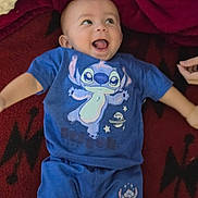 Elias is registered to the contest to win money with this photo: baby, child, blue_clothing, smiling, happy, lying_down, blanket, red_blanket, shorts, tshirt, cartoon_character, stitch, infant, cute, indoor, person, portrait, playful, joy, small_child