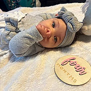 Calissia is registered to the contest to win money with this photo: baby, bed, blanket, bow_headband, child, close_up, cozy, family_photo, fleece, gray_clothing, hand_to_mouth, indoor, infant, lying_down, name_plaque, nursery, portrait, smiling, wide_eyes, wooden_sign