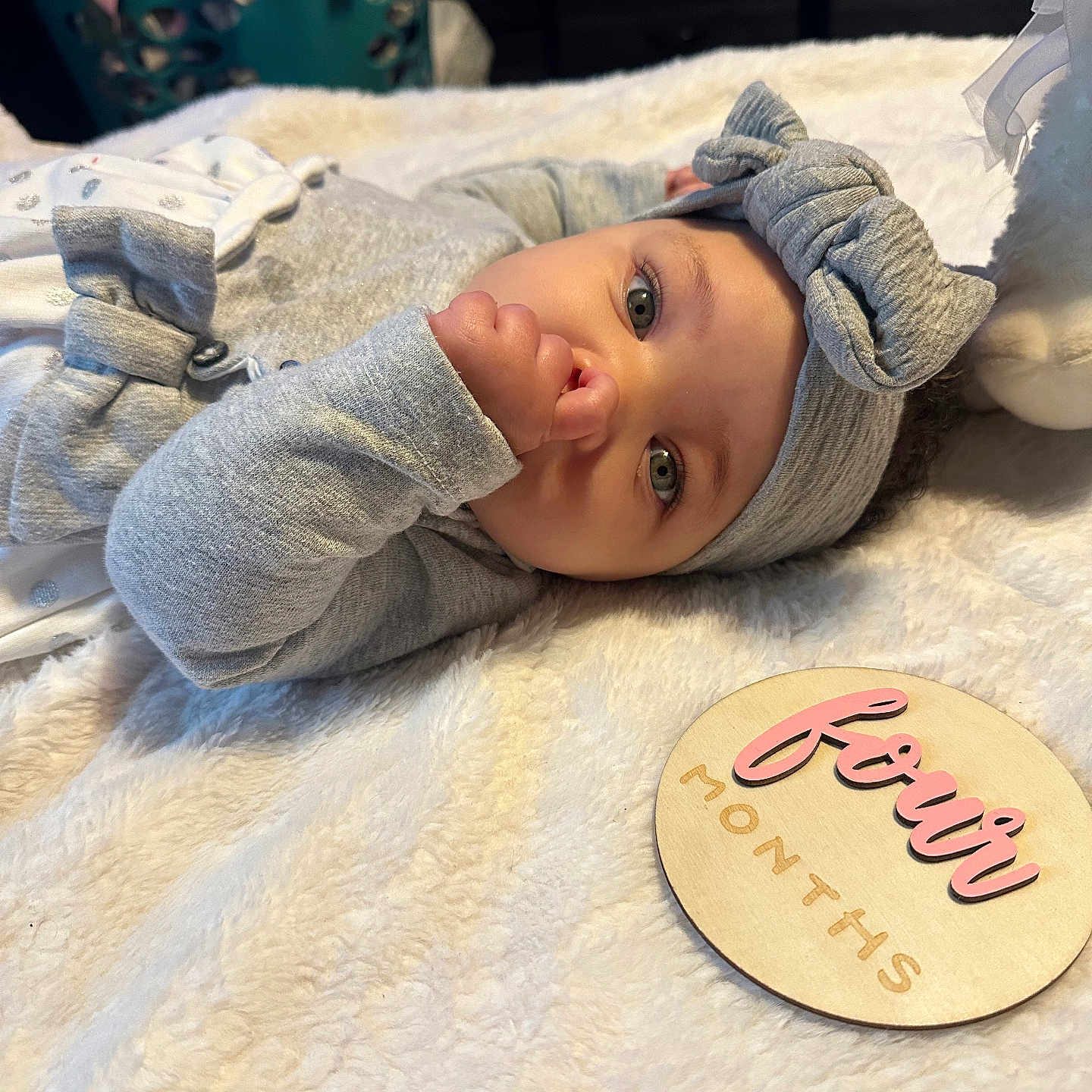 Calissia is registered to the contest to win money with this photo: baby, bed, blanket, bow_headband, child, close_up, cozy, family_photo, fleece, gray_clothing, hand_to_mouth, indoor, infant, lying_down, name_plaque, nursery, portrait, smiling, wide_eyes, wooden_sign