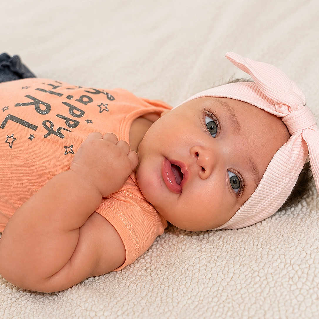 Calissia is registered to the contest to win money with this photo: baby, infant, child, headband, bow, peach_shirt, happy_girl_text, lying_down, soft_blanket, cute, portrait, face, eyes, hand, skin, clothing, indoors, young_child, adorable, expression