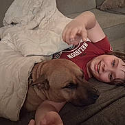 Bane is registered to the contest to win money with this photo: child, dog, blanket, couch, smiling, happy, indoor, relaxing, pet, friendship, comfort, casual, cozy, brown_dog, portrait, home, cute, resting, young_child, friendly