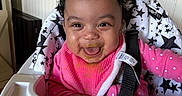 Wynter is registered to the contest to win money with this photo: baby, child, high_chair, pink_clothing, bib, food_smear, curly_hair, smiling, happy, indoor, face, seatbelt, star_pattern, mealtime, kitchen, toddler, cute, messy, expression, person