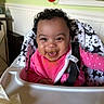 baby, child, high_chair, pink_clothing, bib, food_smear, curly_hair, smiling, happy, indoor, face, seatbelt, star_pattern, mealtime, kitchen, toddler, cute, messy, expression, person