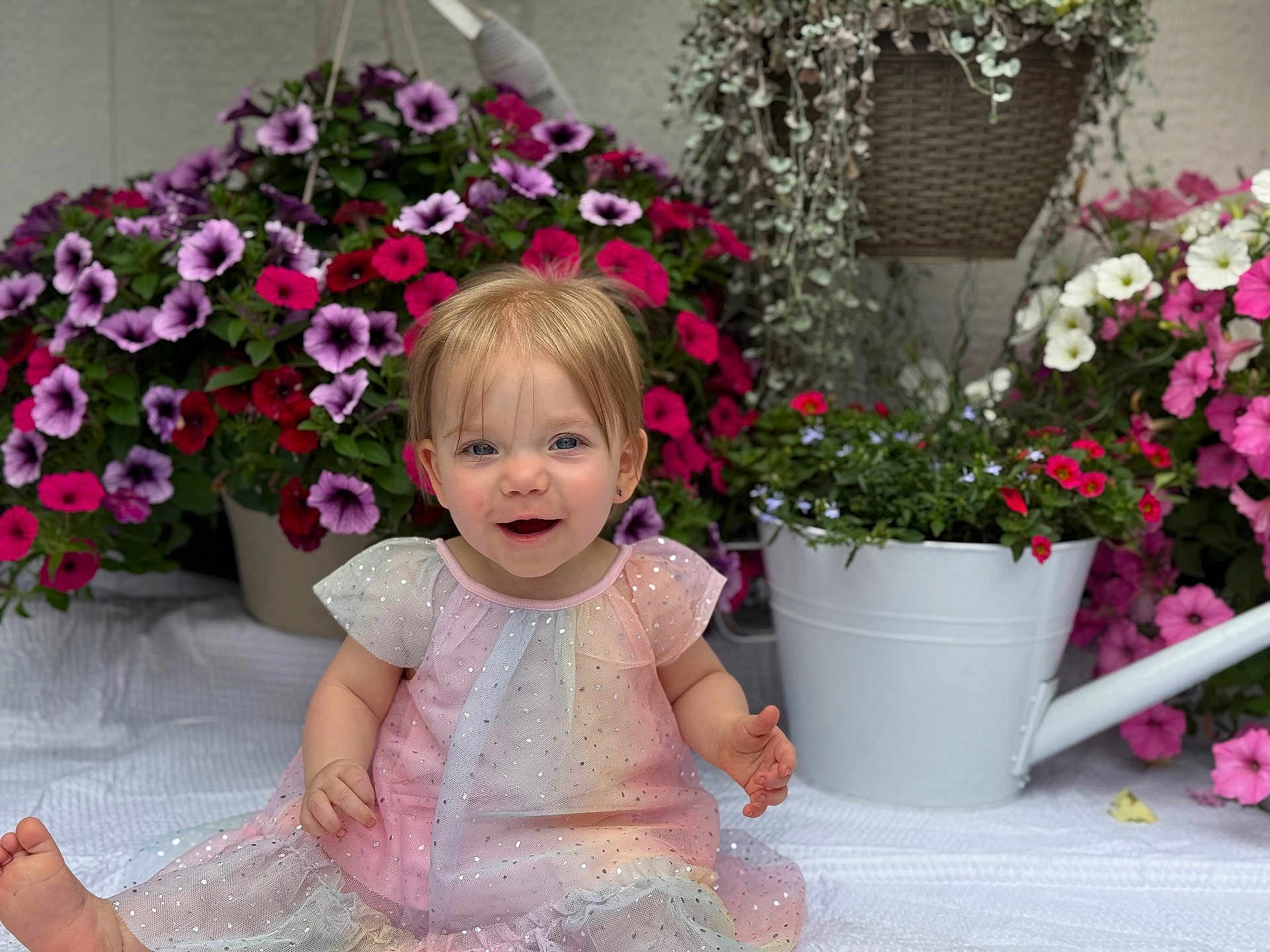 Barbara is registered to the contest to win money with this photo: toddler, child, smiling, dress, flowers, potted_plants, watering_can, blanket, garden, pink, purple, white, greenery, cute, happy, sitting, indoor, portrait, baby, face