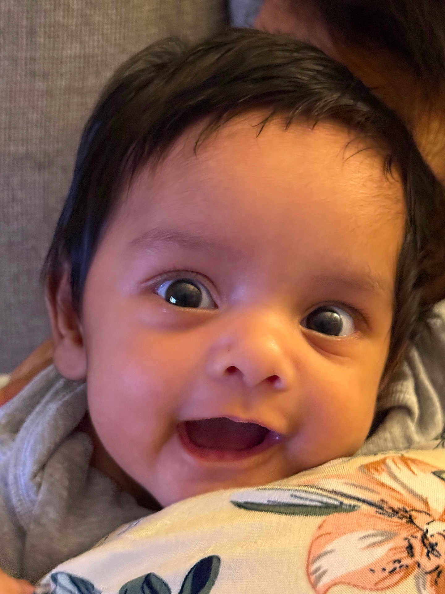 Mehsan is registered to the contest to win money with this photo: baby, infant, face, eyes, smile, expression, dark_hair, close_up, pillow, floral_pattern, soft_background, cute, child, person, indoors, portrait, young, happy, resting, cozy