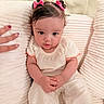 baby, child, infant, pink_bows, hair, cushion, white_clothing, orange_trim, hand, nail_art, soft_texture, seated, indoors, portrait, cute, comfortable, skin, face, person, warmth