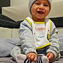 baby, bib, child, clothing, cute, expressive, face, hand, happy, hat, indoors, infant, pom_poms, portrait, seated, smiling, socks, soft_surface, striped_socks, warm