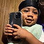 Tamir joined the competition — help win amazing prizes! toddler, child, smiling, holding_phone, black_head_wrap, green_bib, baby, happy, indoor, wood_panel_wall, adult_hand, glasses, close_up, portrait, technology, cute, person, skin, hand, face