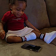 Tamir is registered to the contest to win money with this photo: toddler, child, smiling, sitting, couch, phone, red_shirt, white_shorts, white_shoes, striped_socks, indoor, hair_puffs, happy, casual_clothing, furniture, relaxed, person, home, technology, young_child