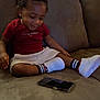toddler, child, smiling, sitting, couch, phone, red_shirt, white_shorts, white_shoes, striped_socks, indoor, hair_puffs, happy, casual_clothing, furniture, relaxed, person, home, technology, young_child