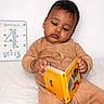 baby, child, infant, reading, book, corduroy, pajamas, milestone_card, 4_months, white_background, sitting, indoors, cute, young, portrait, cozy, hand, face, expression, education