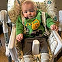 animal_print, baby, child, clothing, constellation_print, cozy, cute, excavator, face, furniture, green_sweater, high_chair, indoor, person, playful, seat_belt, smiling, sock, toddler, wooden_floor