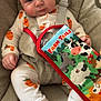 baby, infant, child, carrot_pajamas, baby_seat, straps, farm_tails_book, toy_ring, animal_cartoon, cute, indoors, soft_toy, childrens_book, face, hand, foot, expression, clothing, seat, playtime