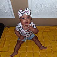 Diamond is registered to the contest to win money with this photo: baby, child, infant, sitting, yellow_surface, headband, bow, heart_pattern, clothing, cute, indoor, curious, hand_gesture, portrait, person, young_child, smiling, playful, casual, fashion