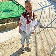 Tiana participe au concours pour gagner de l'argent avec cette photo : child, girl, glasses, shawl, fringe, white_blouse, jeans, sneakers, outdoor, sunlight, shadow, concrete, smile, happy, standing, bracelet, gate, stone_wall, green_cloth, chair